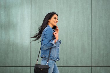 Side portrait of young smiling latin woman walking with mobile phone and purse