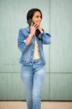 Portrait beautiful young hispanic woman talking with phone while walking by green wall