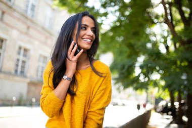 Şehirde cep telefonuyla konuşan gülümseyen genç Latin bir kadın.