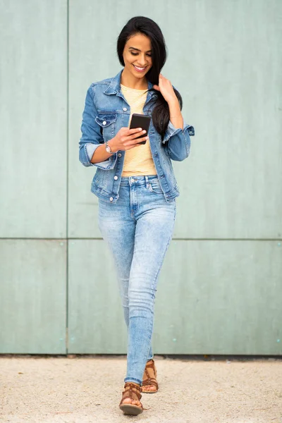 Full length portrait beautiful smiling young latin woman looking at cellphone while walking by green wall