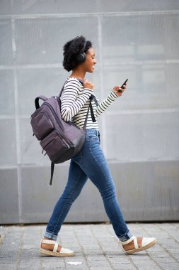 Tam boy portre mutlu siyah kadın gezgin çantayla yürüyor cep telefonu ve kulaklıkla müzik dinliyor. 