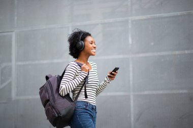Yan portre mutlu genç zenci kadın çantalı gezgin cep telefonu ve kulaklıkla müzik dinliyor. 
