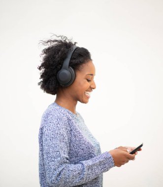 Yandan gülümseyen Afro-Amerikan bir kadın cep telefonu ve kulaklıkla izole edilmiş beyaz arka planda müzik dinliyor.