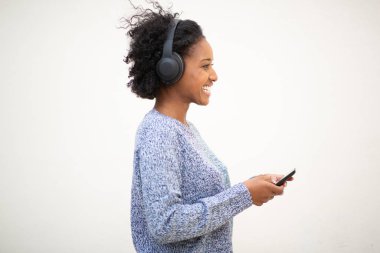 Yandan gülümseyen Afrikalı Amerikalı bir kadın cep telefonu ve kulaklıkla izole edilmiş beyaz arka planda müzik dinliyor.