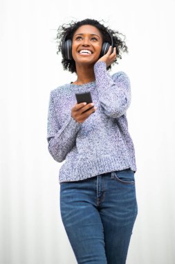 Portre mutlu genç siyah kadın beyaz duvarda cep telefonu ve kulaklıkla müzik dinliyor.