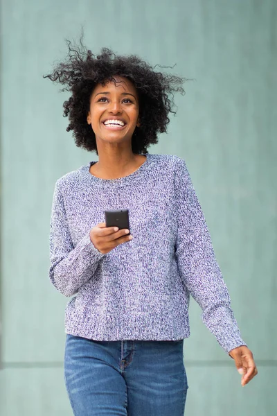 Portrait happy young african American woman walking outside with mobile phone by green wall
