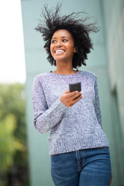 Portrait happy young african American woman walking with cellphone by green wall