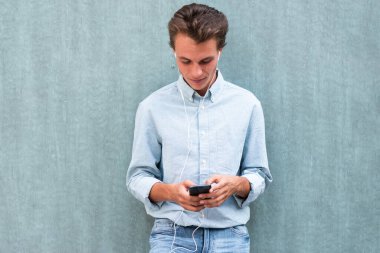 Duvara yaslanmış cep telefonuyla müzik dinleyen genç bir adam portresi.