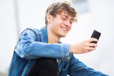 Kapalı portre gülümseyen genç adam dışarıda oturmuş cep telefonuna bakıyor.