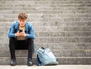 Tam boy portre. Genç adam merdivenlerde oturuyor. Cep telefonuna bakıyor.