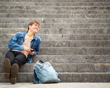Tam boy portre genç adam merdivenlerde rahatlıyor cep telefonunu tutuyor ve başka tarafa bakıyor.