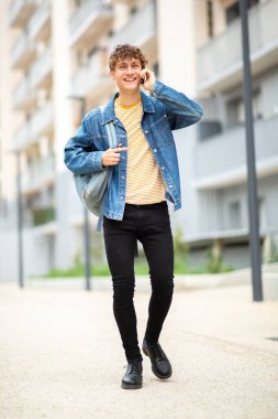 Tam boy portre mutlu genç adam dışarıda telefonla konuşuyor ve yürüyor.