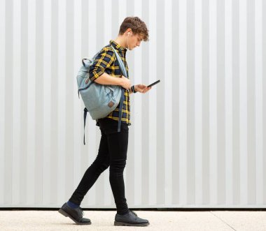 Tam boy yan portre genç adam çantasıyla yürüyor ve beyaz arka planda cep telefonu ve kulaklıkla müzik dinliyor