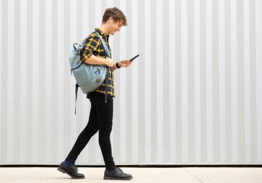 Tam boy yan portre gülümseyen genç adam çantasıyla yürüyor ve beyaz arka planda cep telefonu ve kulaklıkla müzik dinliyor.