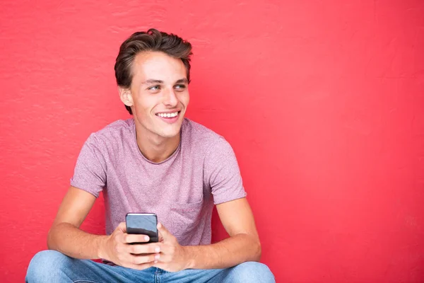 坐在红墙边 拿着手机的年轻人笑着走近肖像 — 图库照片