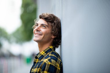 Yan portre gülümseyen genç adam gri duvara yaslanmış yukarı bakıyor ve düşünüyor