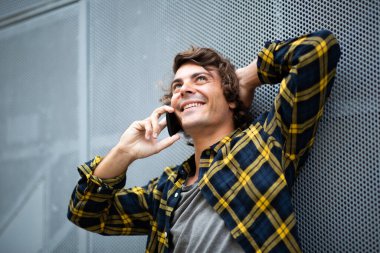 Duvara yaslanmış gülümseyen genç adam cep telefonuyla konuşuyor.