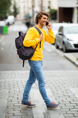 Tam boy profil portresi. Çantalı genç adam cep telefonuyla karşıdan karşıya geçiyor.