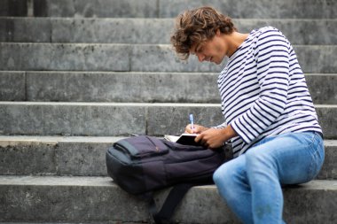 Merdivenlerde oturmuş kitap yazan erkek öğrenci portresi.