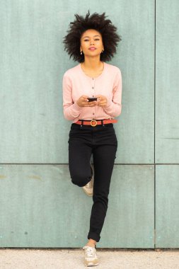 Genç Afrikalı Amerikalı kadın cep telefonuyla duvara yaslanıyor.