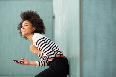 Cep telefonuyla duvara yaslanan mutlu Afro-Amerikan kadın portresi.