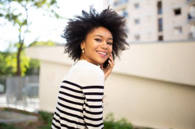 Yandan gülümseyen genç zenci kadın dışarıda cep telefonuyla konuşuyor.