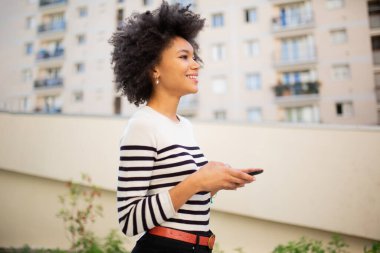 Yandan gülümseyen siyah kadın akıllı telefonuyla dışarıda yürüyor.