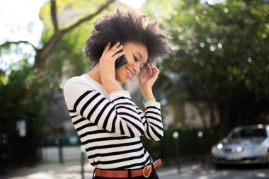 Portre gülümseyen Afrikalı Amerikalı genç bir kadın dışarıda cep telefonuyla konuşuyor.