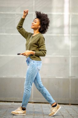 Cep telefonu ve kulaklıklarıyla yürüyen uzun boylu, neşeli, siyah bir kadın.