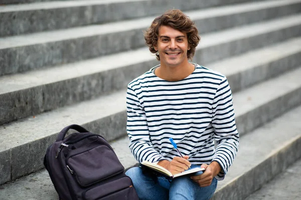Portre Gülümseyen Erkek Üniversite Öğrencisi Dışarıda Oturmuş Kitabı Yazıyor Düşünüyor — Stok fotoğraf