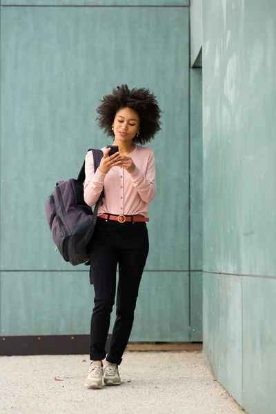 全身ポートレート若いですアフリカ系アメリカ人女性で歩くと携帯電話とバッグによって緑の壁 — ストック写真