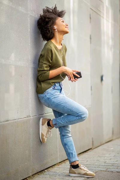 Full body portrait smiling young african woman with mobile phone and earphones leaning against wall
