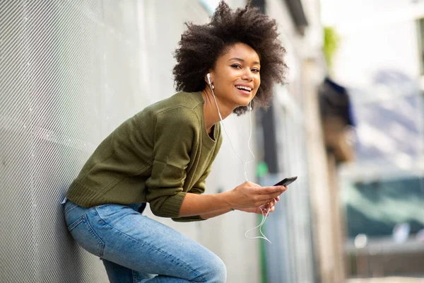 带着手机和耳机靠在墙上的快乐的非洲裔美国女人 — 图库照片