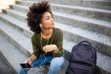 Dışarıda merdivenlerde oturmuş kulaklık ve telefonla müzik dinleyen güzel zenci kadın portresi.