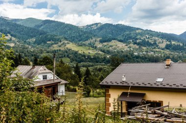 Kahverengi çatılı bir ev mavi gökyüzünün altındaki yeşil ağaçlarla dağlarda duruyor..