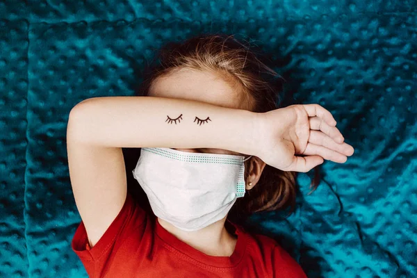 Niño en un vestido rojo se encuentra en un cuadros azules en una máscara médica — Foto de Stock