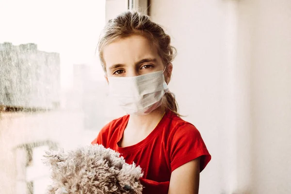 Tıbbi maskeli bir çocuk bir ayıyla pencereye oturur.