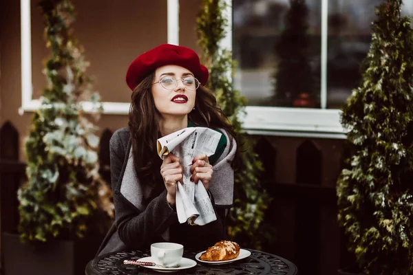 Gözlüklü, gri montlu, kırmızı bereli bir kız elinde bir gazeteyle caddede bir kafede oturmuş kruvasanla kahve içiyor. Bir toplantı. İş kadını. Paris mi? Gösterge