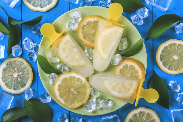 Lezzetli ve ferahlatıcı limonlu dondurma lolipopları mavi ahşap bir masada buz küpleri, limon dilimleri ve doğa yapraklarıyla kaplı. Yaz pikniği.