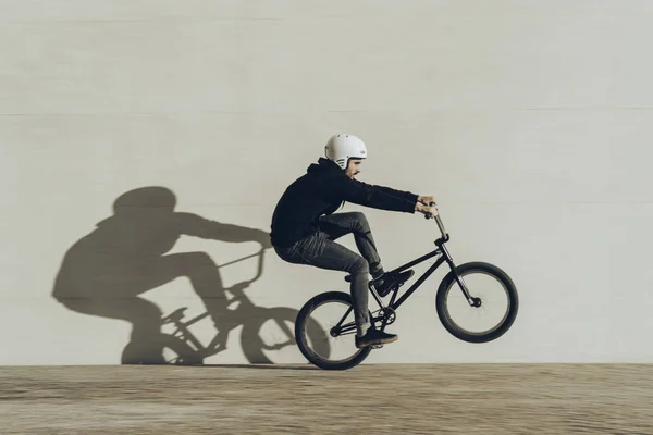 Bmxer doing a wheelie with his shadow projected in a stone wall