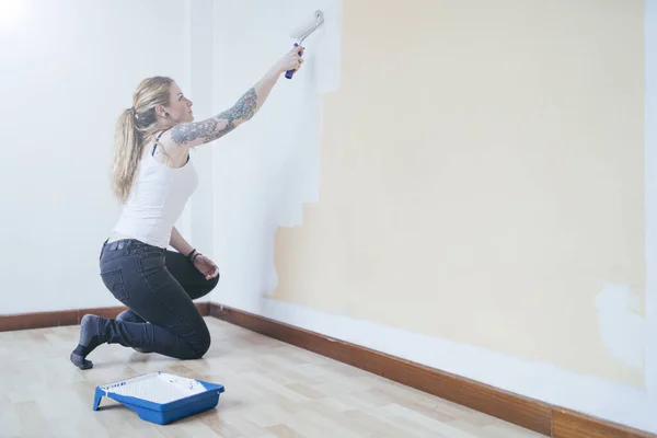 Young blonde inked woman painting a room bent down. She is painting a room with white pain. She is wearing white t-shirt and black jeans.
