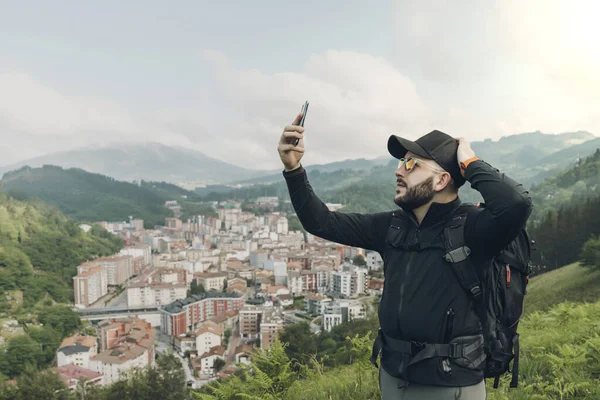 Young man worried with no signal on smartphone. He is holding it on top and head with other hand. Wearing backpack, dark sportswear and cap. Village in valley rounded by mountains and blue sky in back