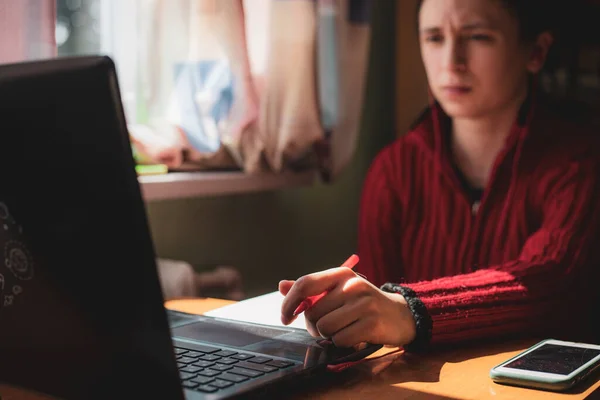 Ung kaukasiska flicka i röd tröja med förvirrad blick på hennes ansikte tittar på datorn när du studerar online hemma — Stockfoto