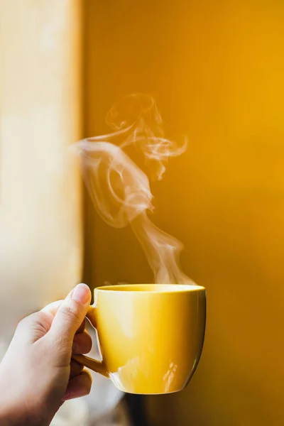 Yellow cup with hot steaming tea drink in hand near window with bright yellow wall background in beautiful morning light