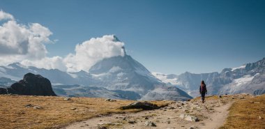 Sırt çantalı bir kız kar kaplı güzel İsviçre Alplerinde iz sürerek yürüyor ve bulutlarla kaplı Matternhorn, parlak mavi gökyüzü
