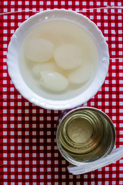 Tasty white canned rambutan in heavy sweet syrop in white bowl near can with bright red background