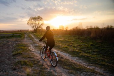 Kırsal alanda Susnet 'e doğru bisiklet süren ceketli ve şapkalı bir kız.