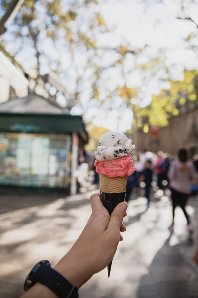 Hand holding cone with colorful delicious gelato ice-cream in touristic La Rambla street in Barcelons in summer day with busy crowded street background