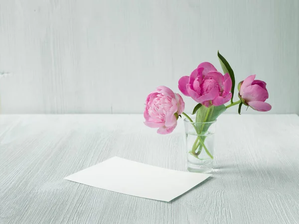 Pfingstrosen Strauß in Klarglasvase. Saubere Dekoration im skandinavischen Stil. Leere Einladungskarte-Attrappe auf weißem Holztisch — Stockfoto