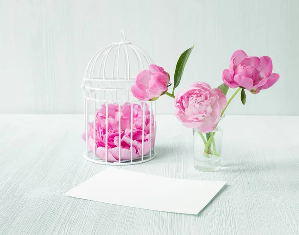 Weiße Vogelkäfige mit Blütenblättern auf einem Holztisch. Drei Pfingstrosen blühen in einer Glasvase. Leere Einladungskarte für die Hochzeitsfeier. Saubere Dekoration im skandinavischen Stil — Stockfoto